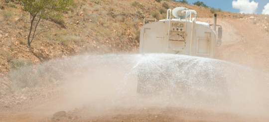 Water truck driving away from the viewer spraying water on a dirt road for heavy equipment