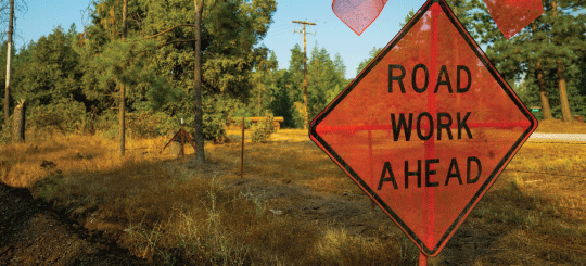 Orange Road Work Ahead sign on a rural road in a natural area with trees and grasses in California