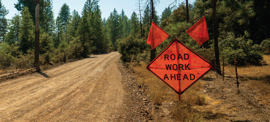 Orange road construction signs on a forested, unpaved, Perma-Zyme treated road