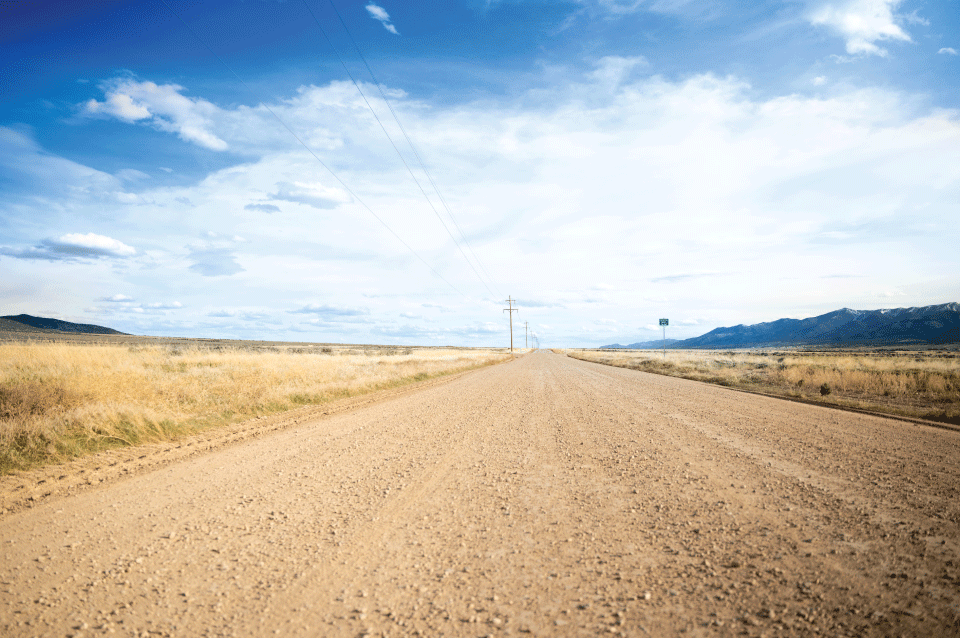 Low-angle photo of an unpaved, Perma-Zyme treated road in a rural grassland with mountains and utility poles in the background