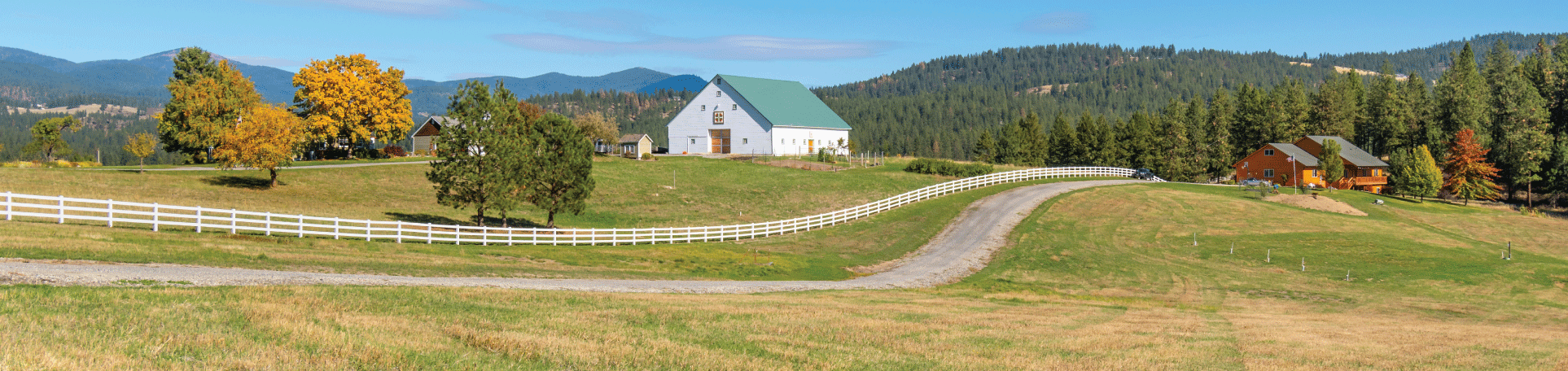 Residential farm property with driveway, log house, white barn, white fence, and mountains in the background